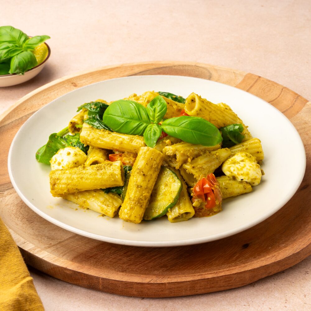 Op deze afbeelding is een bruine houten plank zichtbaar, met daarop een wit bord met het gerecht. Het gerecht bestaat uit rigatoni pasta met Pesto alla Genovese, courgette, tomaatjes en mozzarella. Het gerecht is gegarneerd met een mooi blaadje basilicum bovenop.