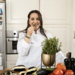 Een foto van Hilal, ook wel bekend als Moon's kitchen. Ze draagt een witte chef-jas en ruikt aan een bosje verse kruiden. Op de tafel voor haar liggen verse groenten, zoals aubergine, courgette en tomaat. Hilal glimlacht en ze heeft een sprankeling in haar bruine ogen.