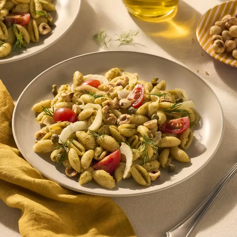 Een heerlijk bordje vol geribbelde schelpjespasta, omhult met groene pesto alla genovese. Tussen de pastaschelpjes zitten tomaatjes en stukjes venkel. De tafel is gedekt met een geel servet en nog wat hazelnootjes als garnering in een geel gestreept bordje.