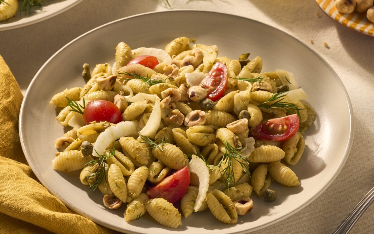 Een heerlijk bordje vol geribbelde schelpjespasta, omhult met groene pesto alla genovese. Tussen de pastaschelpjes zitten tomaatjes en stukjes venkel. De tafel is gedekt met een geel servet en nog wat hazelnootjes als garnering in een geel gestreept bordje.