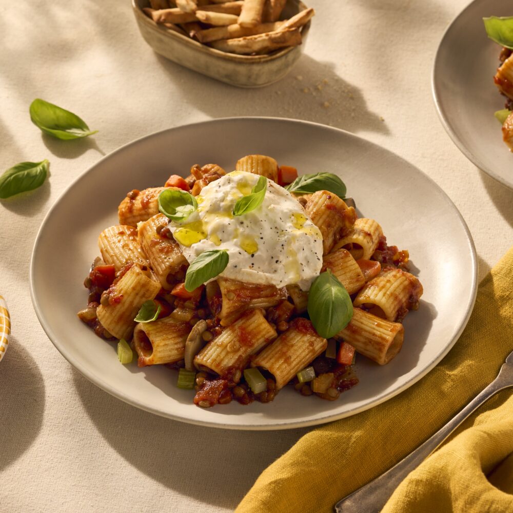 Een heerlijk bordje pasta met halve rigatoni. De geribbelde buisjespasta houdt de vegetarische bolognesesaus (gemaakt van linzen) goed vast en dat zie je goed op de afbeelding. Het gerecht is gegarneerd met wat verse basilicum blaadjes en bovenop ligt een heerlijke burrata.