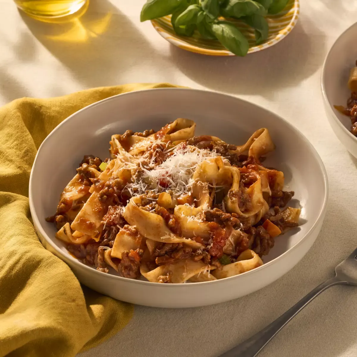 Op een mooi gedekte tafel staat een bord pappardelle pasta. De brede pasta linten worden omhult door een ragu (gemaakt op een snelle manier met gehakt). Je ziet de stukjes groente en vlees in de saus goed zitten. Het gerecht is gegarneerd met geraspte Parmezaanse kaas.