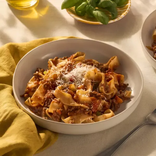 Op een mooi gedekte tafel staat een bord pappardelle pasta. De brede pasta linten worden omhult door een ragu (gemaakt op een snelle manier met gehakt). Je ziet de stukjes groente en vlees in de saus goed zitten. Het gerecht is gegarneerd met geraspte Parmezaanse kaas.