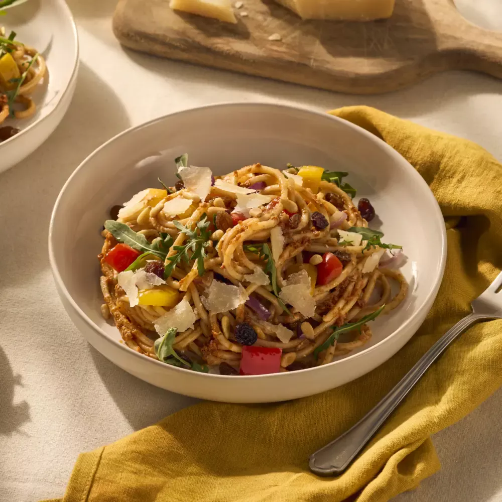 Op het bord ligt de vierkante spaghetti, die lekker wordt omhult door de rode pesto. Tussen de pastaslierten zijn de gele en rode paprika goed zichtbaar, ook zie je wat rucola en rozijnen in het gerecht. Als garnering is het gerecht afgemaakt met Parmezaanse kaas flakes.