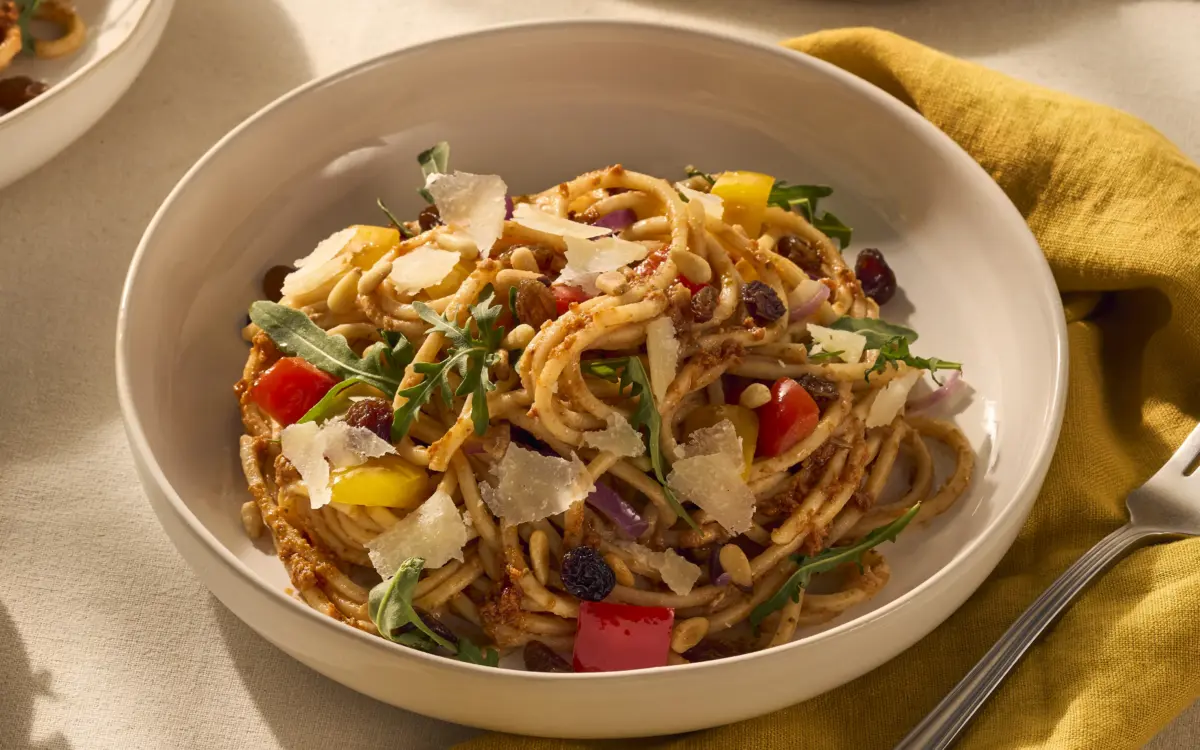 Op het bord ligt de vierkante spaghetti, die lekker wordt omhult door de rode pesto. Tussen de pastaslierten zijn de gele en rode paprika goed zichtbaar, ook zie je wat rucola en rozijnen in het gerecht. Als garnering is het gerecht afgemaakt met Parmezaanse kaas flakes.