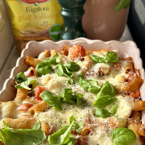 Een ovenschotel in een ovenschaal met golvend randje. De ovenschotel is gemaakt met rigatoni pasta en heeft een heerlijke topping van gesmolten kaas en basilicumblaadjes. Op de achtergrond staat de verpakking Grand'Italia Rigatoni Half Volkoren in beeld.