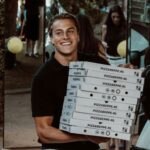 Wisse, een leuke spontane jongen met een glimlach op zijn gezicht. Wisse heeft blond haar en draagt een zwarte polo. In zijn handen heeft hij een stapel pizza dozen. 