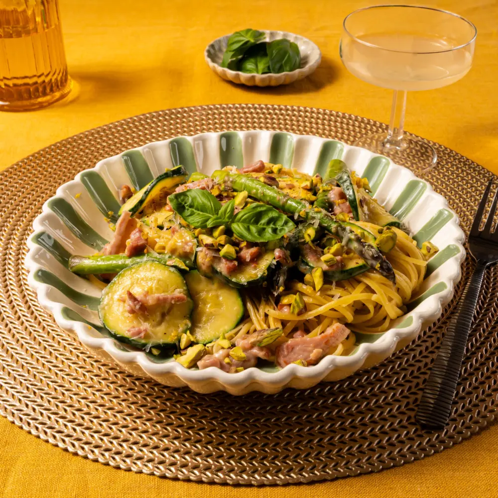 En vrolijk gekleurd bord met groene streepjes. Op het bord ligt het recept van linguine met groene asperges en pancetta. Er ligt een blaadje basilicum bovenop.