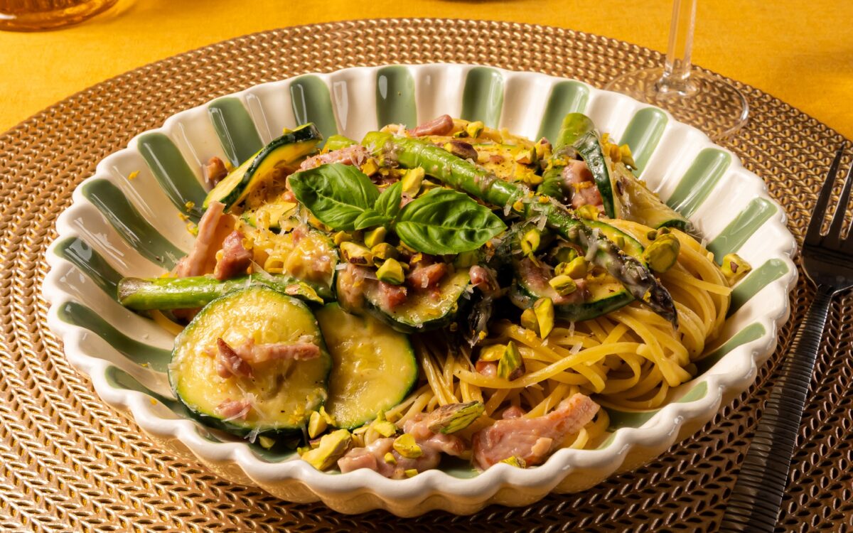 En vrolijk gekleurd bord met groene streepjes. Op het bord ligt het recept van linguine met groene asperges en pancetta. Er ligt een blaadje basilicum bovenop.
