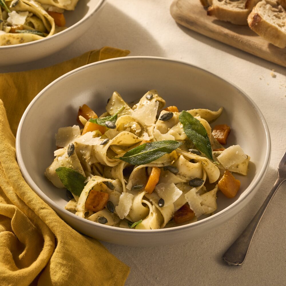 Op een gedekte tafel staat een bord met pappardelle. Het gerecht is aangekleed met geroosterde stukjes oranje pompoen en knapperige groene salieblaadjes. Naast het bord ligt een vork en een geel servet.