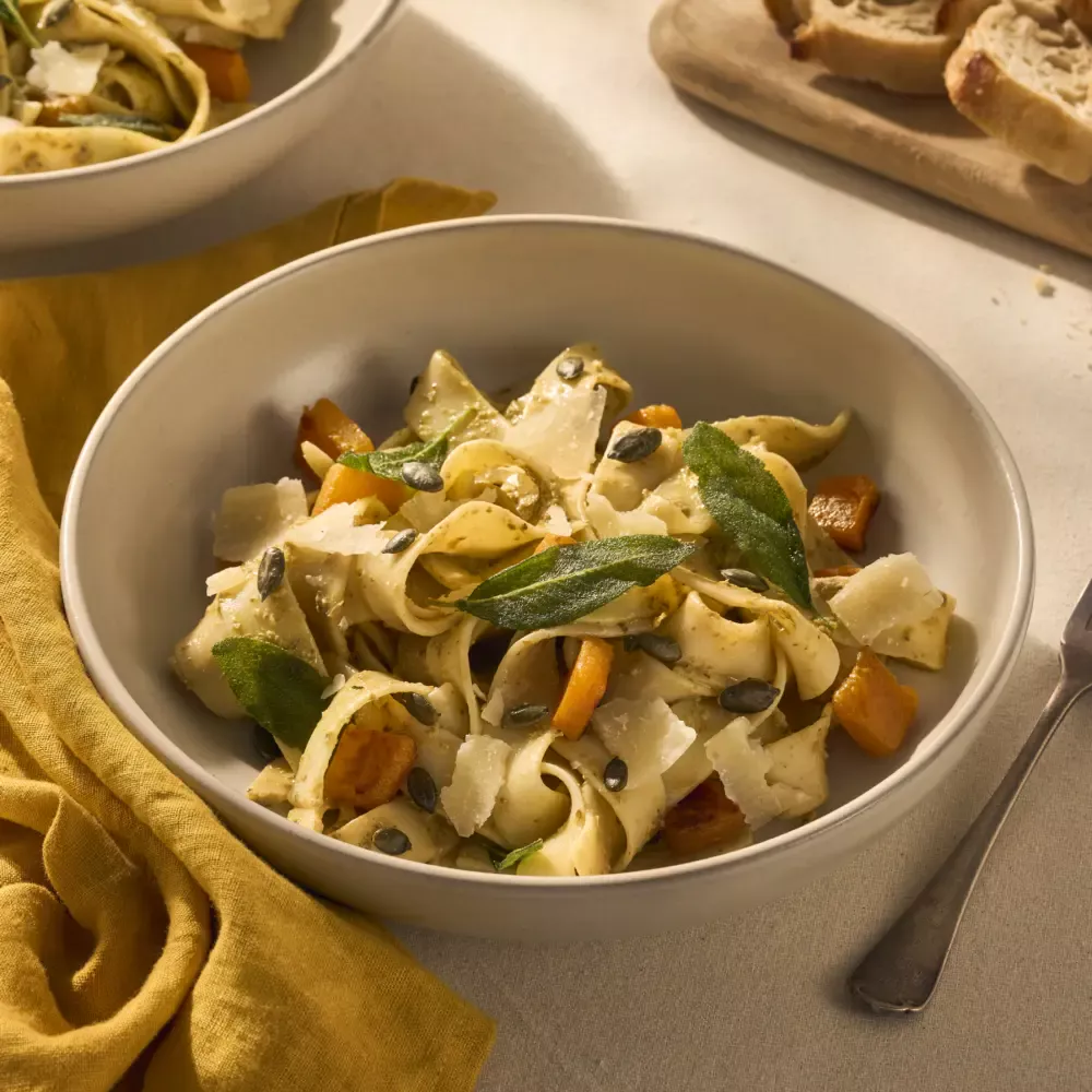 Op een gedekte tafel staat een bord met pappardelle. Het gerecht is aangekleed met geroosterde stukjes oranje pompoen en knapperige groene salieblaadjes. Naast het bord ligt een vork en een geel servet.