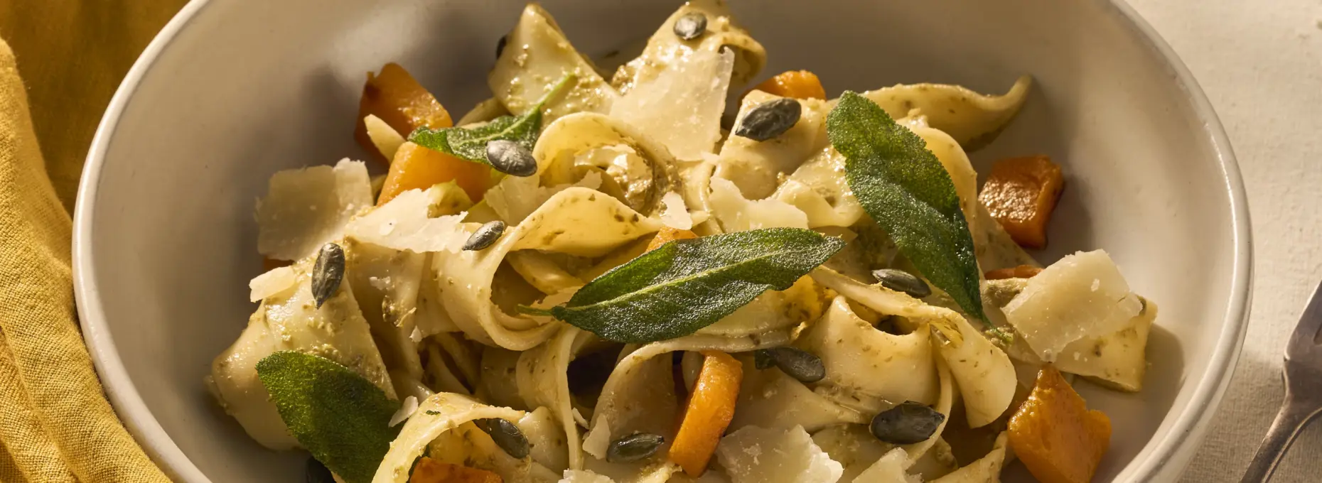 Op een gedekte tafel staat een bord met pappardelle. Het gerecht is aangekleed met geroosterde stukjes oranje pompoen en knapperige groene salieblaadjes. Naast het bord ligt een vork en een geel servet.