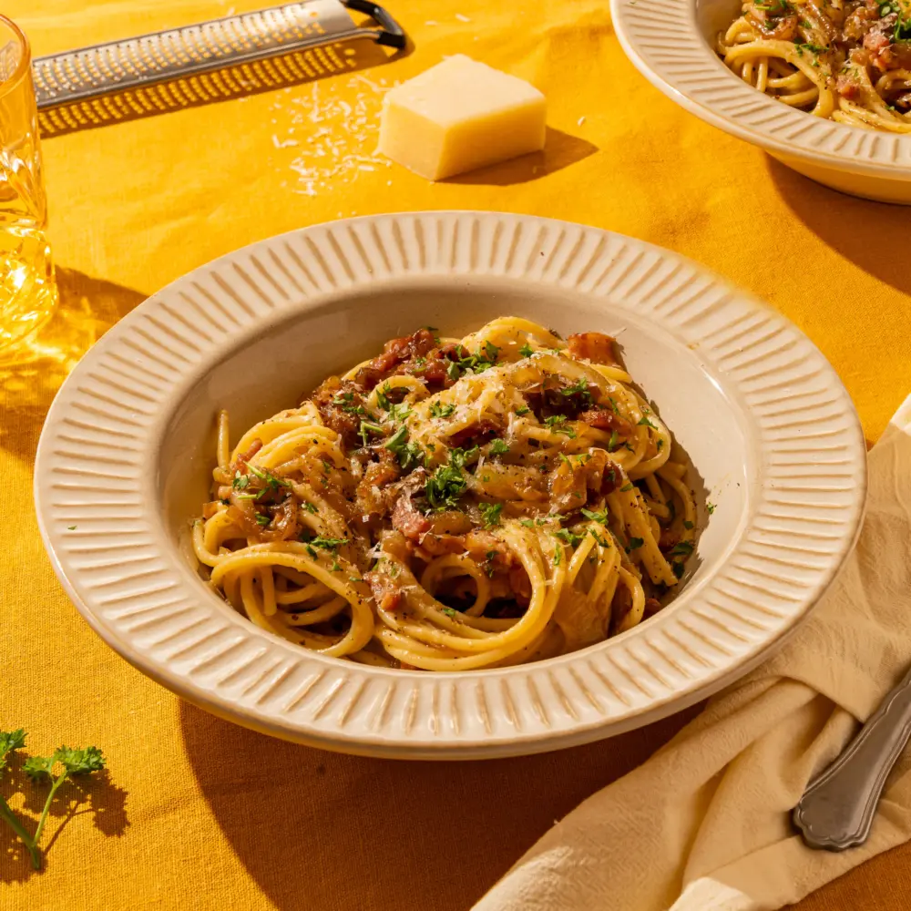 Een heerlijk bord met pasta carbonara. Je ziet tussen de spaghetti slierten de gekaramelliseerde ui zitten en de spekjblokjes. Het gerecht is gegarneerd met een paar stukje peterselie. Naast het bord ligt een rasp met Parmezaanse kaas, waarmee het gerecht extra romig gemaakt kan worden.