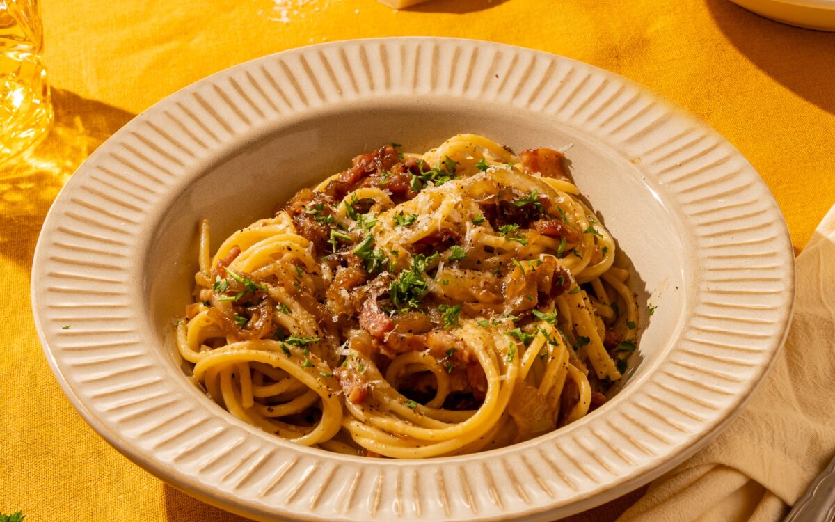 Een heerlijk bord met pasta carbonara. Je ziet tussen de spaghetti slierten de gekaramelliseerde ui zitten en de spekjblokjes. Het gerecht is gegarneerd met een paar stukje peterselie. Naast het bord ligt een rasp met Parmezaanse kaas, waarmee het gerecht extra romig gemaakt kan worden.