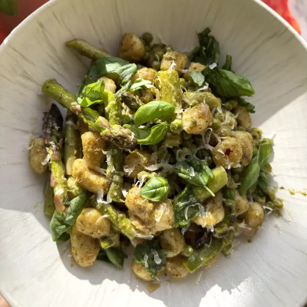 Een heerlijk lente gerecht op een wit bord, waarop zonlicht door het raam valt. In het gerecht zie je de gnocchi, groene asperges en groene pesto goed zitten.