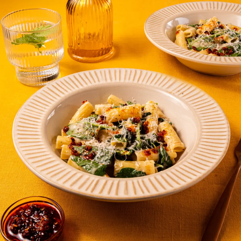 Een bord heerlijk pittige pasta, met rigatoni en spinazie. De romige Romana pastasaus omhult de holle rigatoni en het gerecht is afgemaakt met knapperige crispy chili olie en bestrooid met geraspte Parmezaanse kaas. Op de achtergrond staat een tweede bord en een bakje extra crispy chili olie.