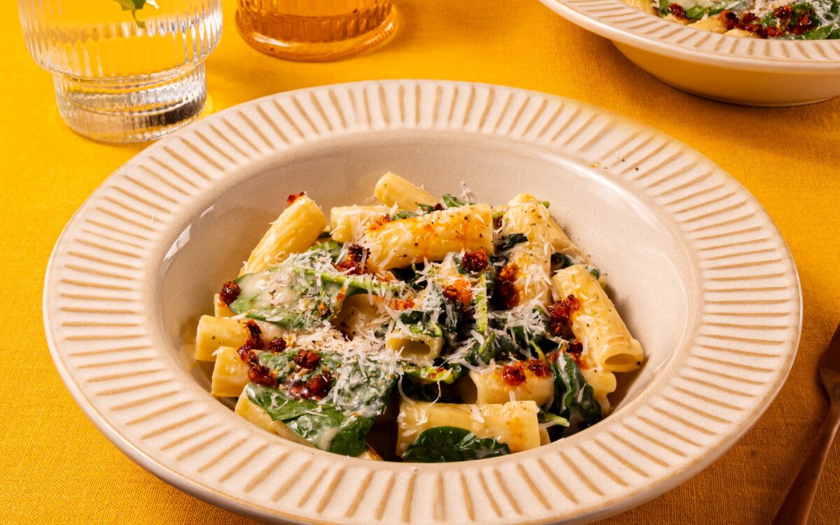 Een bord heerlijk pittige pasta, met rigatoni en spinazie. De romige Romana pastasaus omhult de holle rigatoni en het gerecht is afgemaakt met knapperige crispy chili olie en bestrooid met geraspte Parmezaanse kaas. Op de achtergrond staat een tweede bord en een bakje extra crispy chili olie.