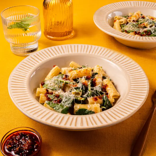 Een bord heerlijk pittige pasta, met rigatoni en spinazie. De romige Romana pastasaus omhult de holle rigatoni en het gerecht is afgemaakt met knapperige crispy chili olie en bestrooid met geraspte Parmezaanse kaas. Op de achtergrond staat een tweede bord en een bakje extra crispy chili olie.