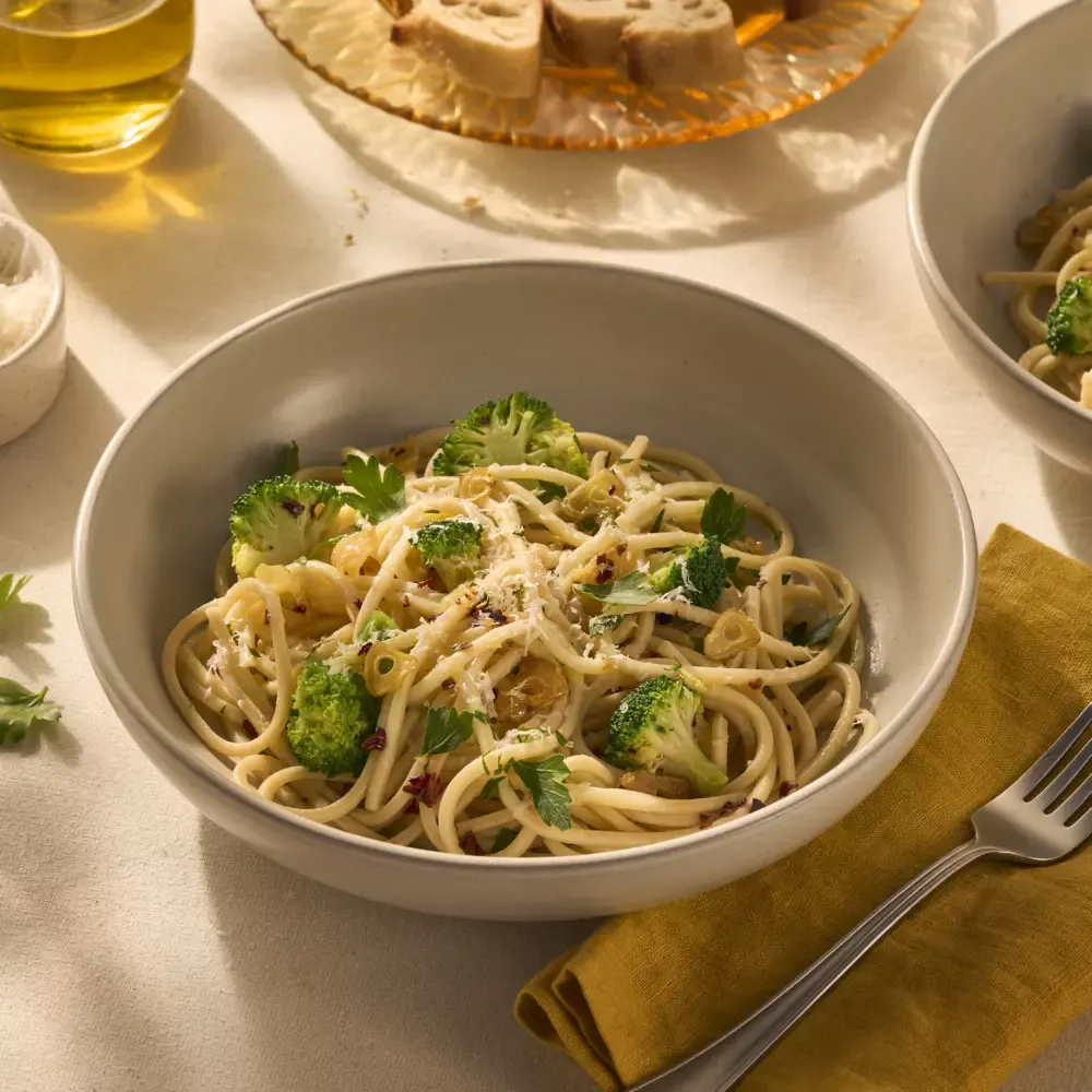 Op de afbeelding zie je een bord pasta met chitarra (een soort vierkante spaghetti), omhult met olijfolie, chilivlokken en knoflook. Tussen de pasta zitten groene verse roosjes broccoli en het gerecht is getopt met geraspte Parmezaanse kaas.