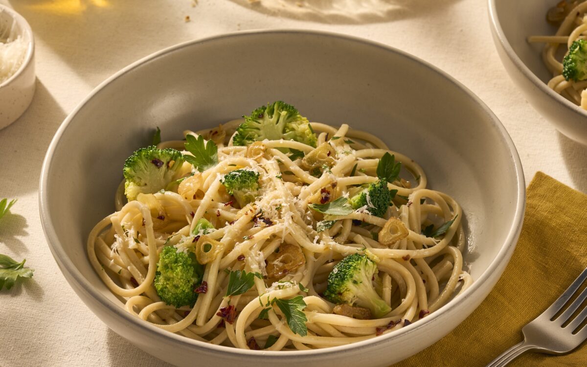 Op de afbeelding zie je een bord pasta met chitarra (een soort vierkante spaghetti), omhult met olijfolie, chilivlokken en knoflook. Tussen de pasta zitten groene verse roosjes broccoli en het gerecht is getopt met geraspte Parmezaanse kaas.