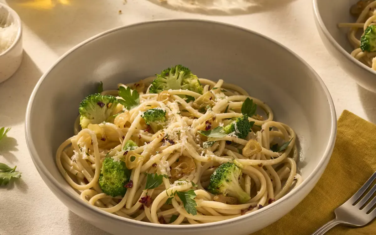 Op de afbeelding zie je een bord pasta met chitarra (een soort vierkante spaghetti), omhult met olijfolie, chilivlokken en knoflook. Tussen de pasta zitten groene verse roosjes broccoli en het gerecht is getopt met geraspte Parmezaanse kaas.