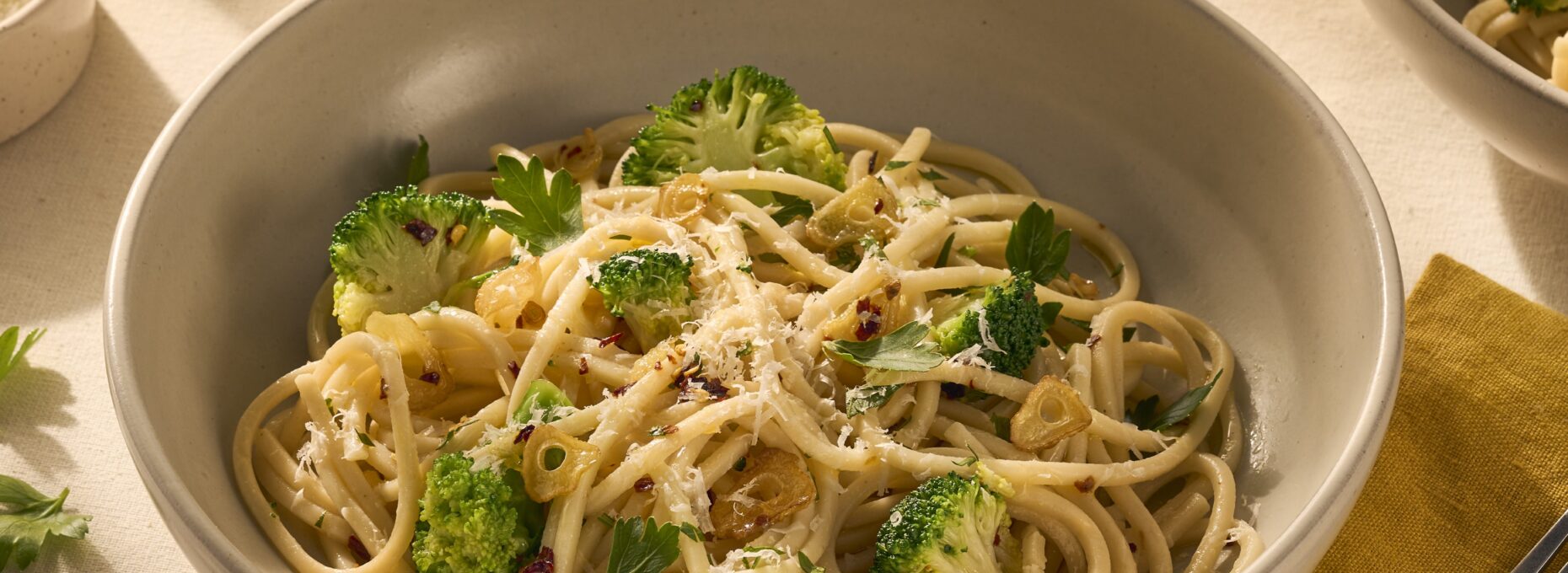 Op de afbeelding zie je een bord pasta met chitarra (een soort vierkante spaghetti), omhult met olijfolie, chilivlokken en knoflook. Tussen de pasta zitten groene verse roosjes broccoli en het gerecht is getopt met geraspte Parmezaanse kaas.