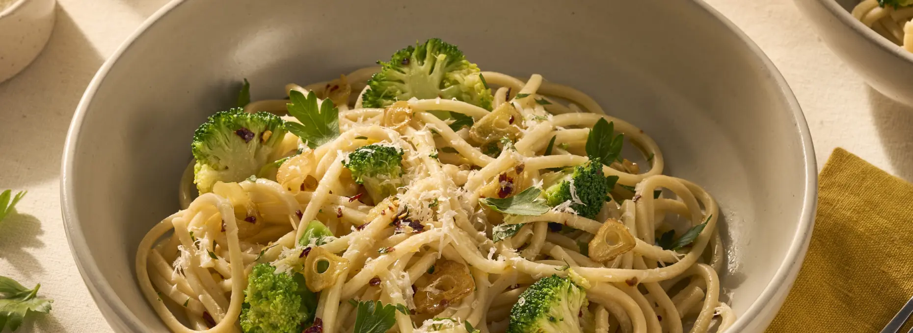 Op de afbeelding zie je een bord pasta met chitarra (een soort vierkante spaghetti), omhult met olijfolie, chilivlokken en knoflook. Tussen de pasta zitten groene verse roosjes broccoli en het gerecht is getopt met geraspte Parmezaanse kaas.