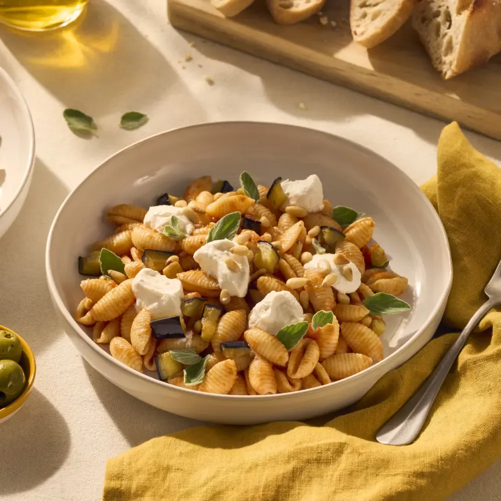 Gnocchetti Sardi zijn kleine Sardijnse pastaschelpjes, die je op deze afbeelding goed kunt zien. In dit gerecht is onze basilico tomatensaus gebruikt en je ziet dat de ribbels en de opening in de schelpjes pasta zich goed vult met de saus. Bovenop de pasta liggen wat dotjes ricotta om het gerecht romiger te maken. Tussen de pasta en de saus zie je de aubergine en de verse blaadjes basilicum zitten. Een heerlijk zonnetje schijnt op tafel, waardoor je je gelijk op het Sardijnse eiland waant.