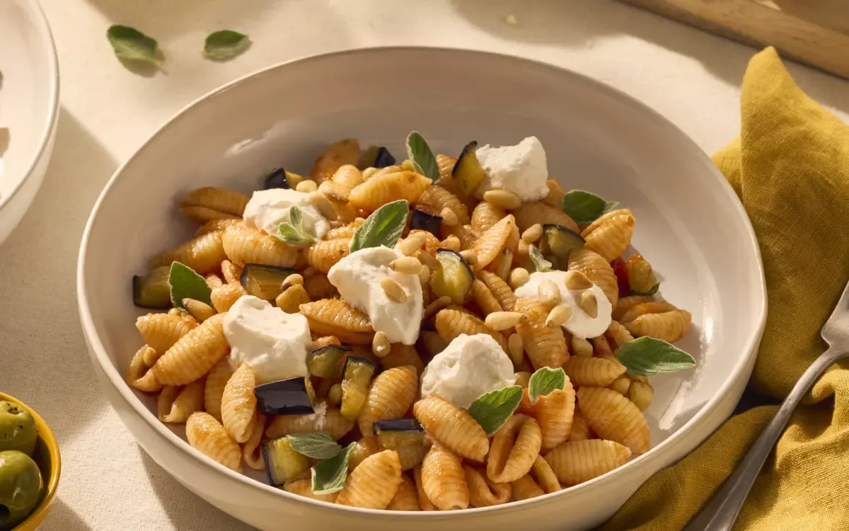 Gnocchetti Sardi zijn kleine Sardijnse pastaschelpjes, die je op deze afbeelding goed kunt zien. In dit gerecht is onze basilico tomatensaus gebruikt en je ziet dat de ribbels en de opening in de schelpjes pasta zich goed vult met de saus. Bovenop de pasta liggen wat dotjes ricotta om het gerecht romiger te maken. Tussen de pasta en de saus zie je de aubergine en de verse blaadjes basilicum zitten. Een heerlijk zonnetje schijnt op tafel, waardoor je je gelijk op het Sardijnse eiland waant.