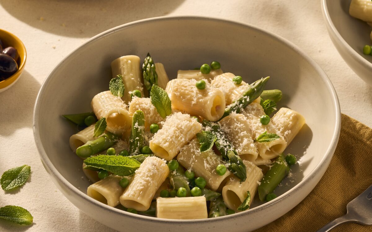 Je ziet een lekker bordje pasta, met kortere rigatoni (geribbelde holle buisjespasta) met een lichte romige carbonara saus. Tussen de pasta en de saus steken groene asperges uit, en bovenop de pasta ligt vers geraspte Parmezaanse kaas.