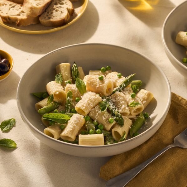 Je ziet een lekker bordje pasta, met kortere rigatoni (geribbelde holle buisjespasta) met een lichte romige carbonara saus. Tussen de pasta en de saus steken groene asperges uit, en bovenop de pasta ligt vers geraspte Parmezaanse kaas.